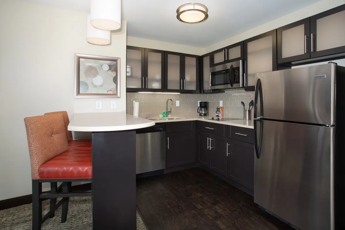 A stylish modern kitchen featuring bar seating, stainless steel appliances, dark cabinetry, and a sleek light countertop. The area is set up for both cooking and casual dining, creating a cozy yet contemporary atmosphere perfect for socializing or enjoying meals.