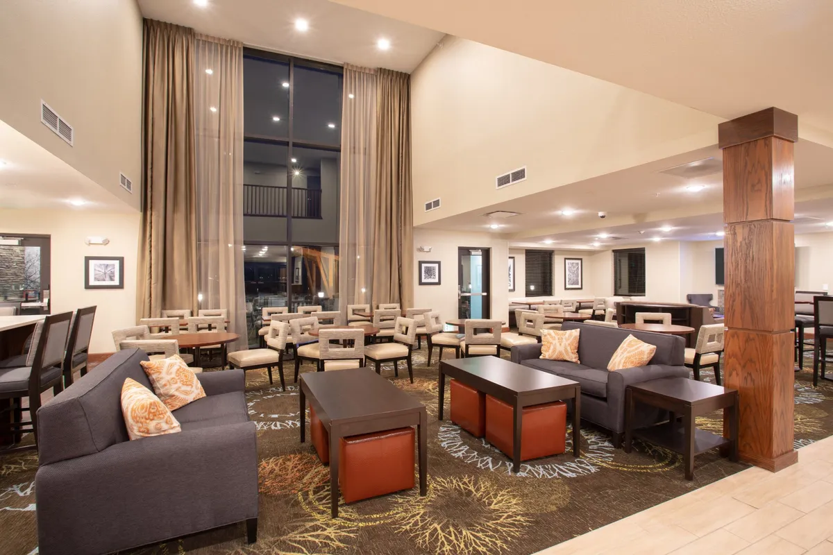 A cozy living room area with large windows that bathe the space in natural light. Plush, comfortable seating surrounds stylish tables, while decorative curtains add a homey touch. The space is arranged to encourage socializing, reading, or relaxing, with an inviting atmosphere ideal for gatherings.