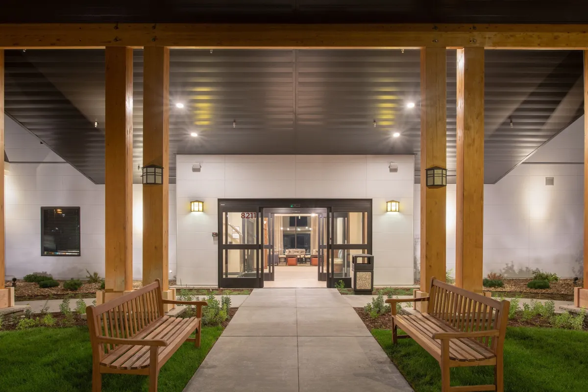 A warmly lit exterior entrance area featuring wooden benches, decorative wall sconces, and well-maintained landscaping. The scene conveys an inviting and welcoming atmosphere, suggesting a pleasant return after a day spent exploring.
