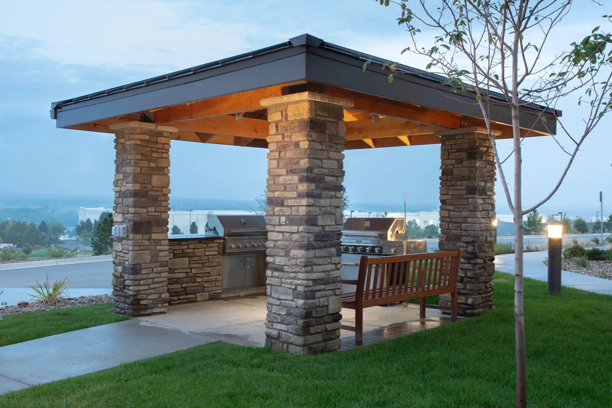 This exterior image showcases a welcoming outdoor area with stone pillars, grill stations, and a wooden bench. The setting is perfect for sunset cookouts, starlit dinners, and informal gatherings, offering guests a space to unwind and network outside of work hours.