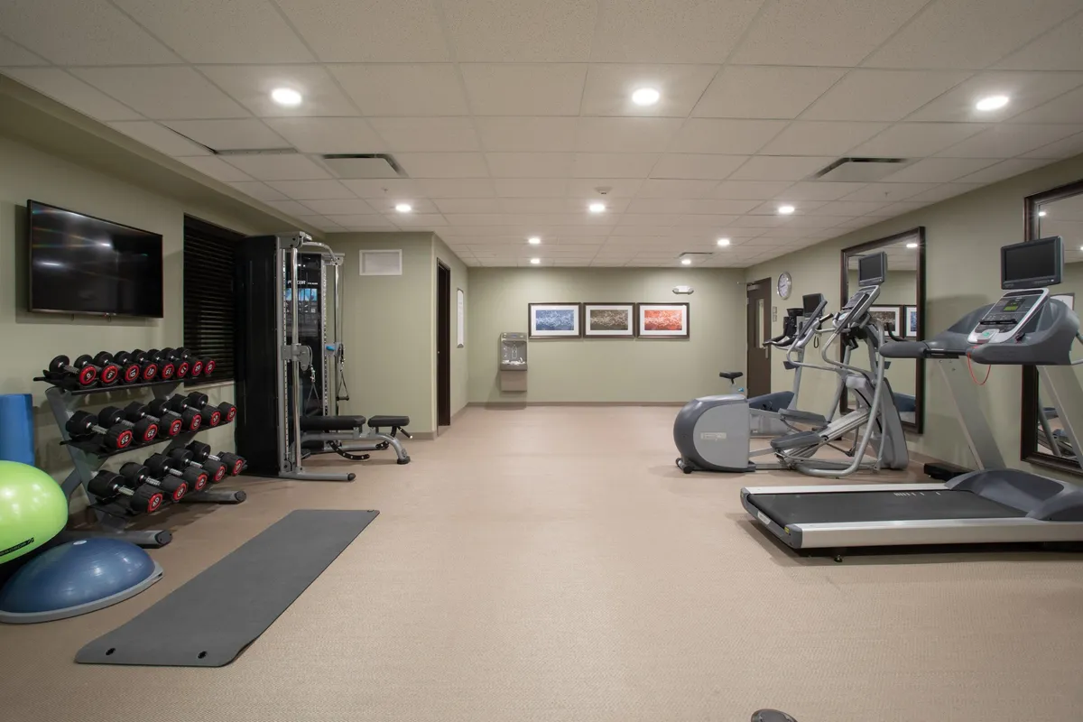 A well-equipped fitness center featuring a rack of dumbbells, treadmills, elliptical machines, an exercise mat, a water cooler, and framed artwork on the walls. The space feels energetic and motivating, ideal for guests wishing to maintain their workout routines during their stay.