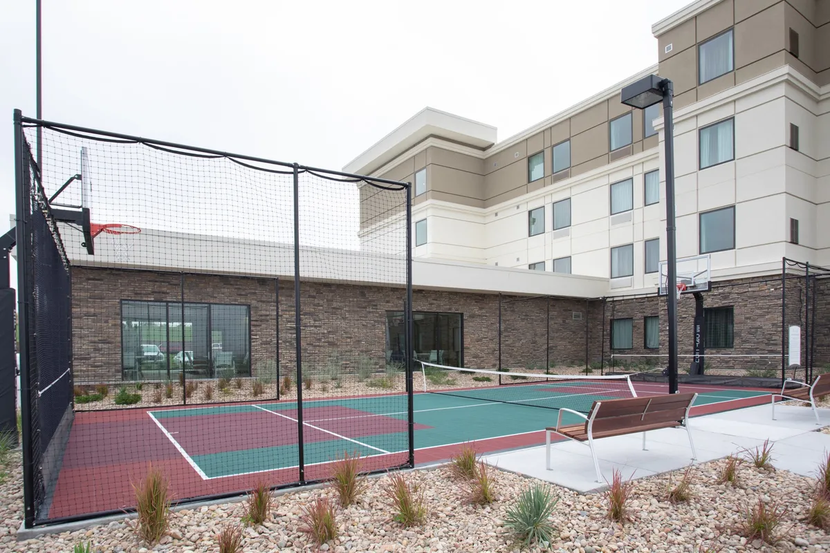 An active outdoor recreation area with a basketball hoop, tennis court, benches, and landscaped surroundings, inviting families to engage in friendly competitions or casual hangouts. The open sky and greenery reinforce the sense of outdoor adventure.